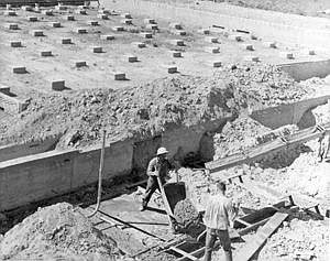 <p>Aug. 22, 1963: With the classroom section of the school well along, work is now beginning on the gymnasium. Here workers pour concrete for the stairway at the northwest gym entrance. In the background are some of the 80 footings which will support the gym floor area.</p>