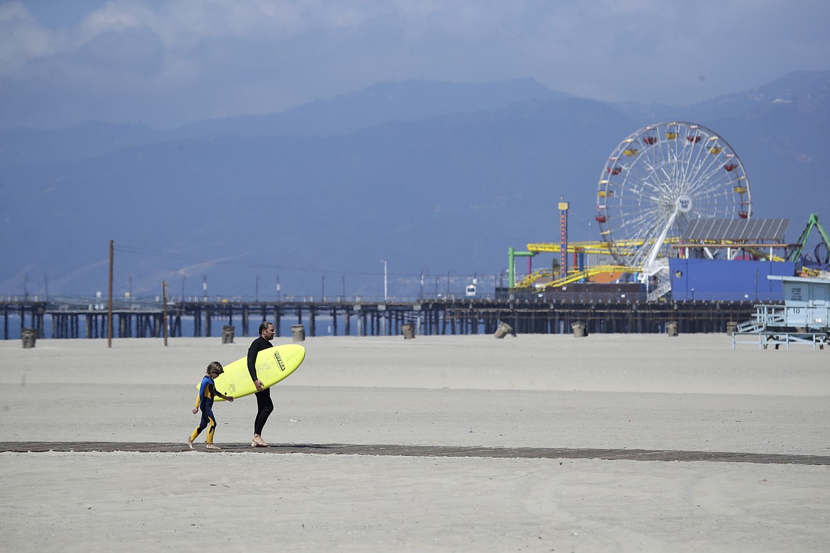 A surfer with a child walks on the mostly empty Santa Monica beach Sunday, March 29, 2020, in Los Angeles. With cases of coronavirus surging and the death toll increasing, lawmakers are pleading with cooped-up Californians to spend a second weekend at home to slow the spread of the infections. (AP Photo/Marcio Jose Sanchez)