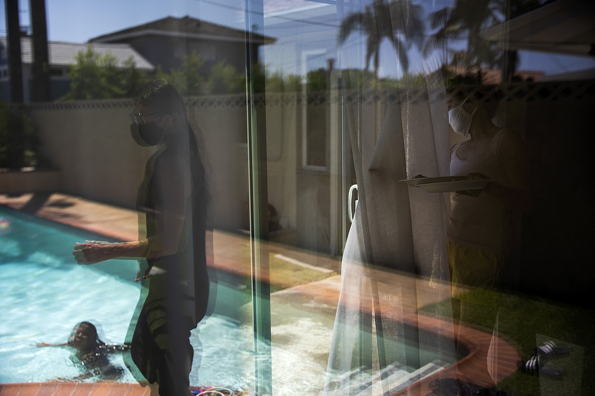 Nurse Jackie Vargas, left, is reflected in a glass door as she walks to her mother, Rose Matute, to pick up lunch for her children playing in their grandmother's pool in Lakewood, Calif., Saturday, July 11, 2020. The 39-year-old nurse, who works in St. Jude Medical Center's COVID-19 unit, doesn't hug her mother-in-law, who is taking care of her children. Through a glass door, she visits her own mom. (AP Photo/Jae C. Hong)