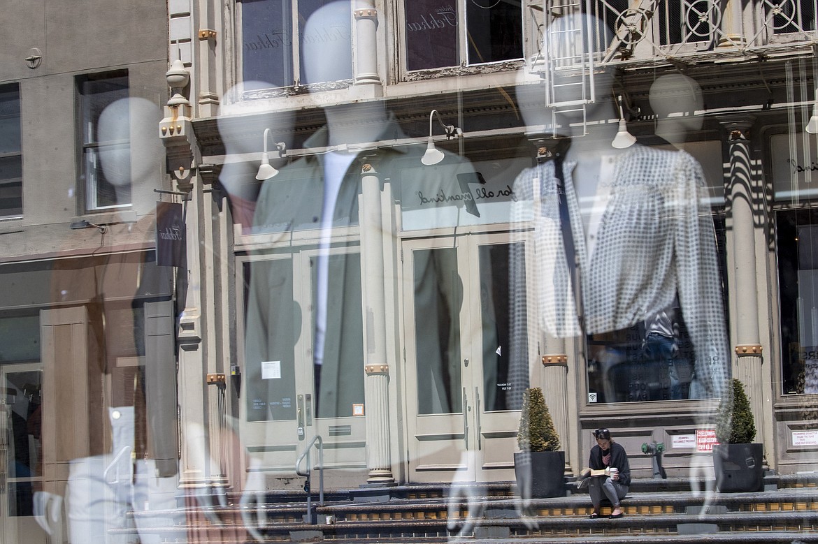 A woman sitting on a stoop reading a book in the sun is seen reflected by a closed clothing store's window on West Broadway, Thursday, May 7, 2020, in the SoHo neighborhood of the Manhattan borough in New York. The U.S. government is poised to report the worst set of job numbers since record-keeping began in 1948, a stunning snapshot of the toll the coronavirus has taken on a now-shattered economy. (AP Photo/Mary Altaffer)