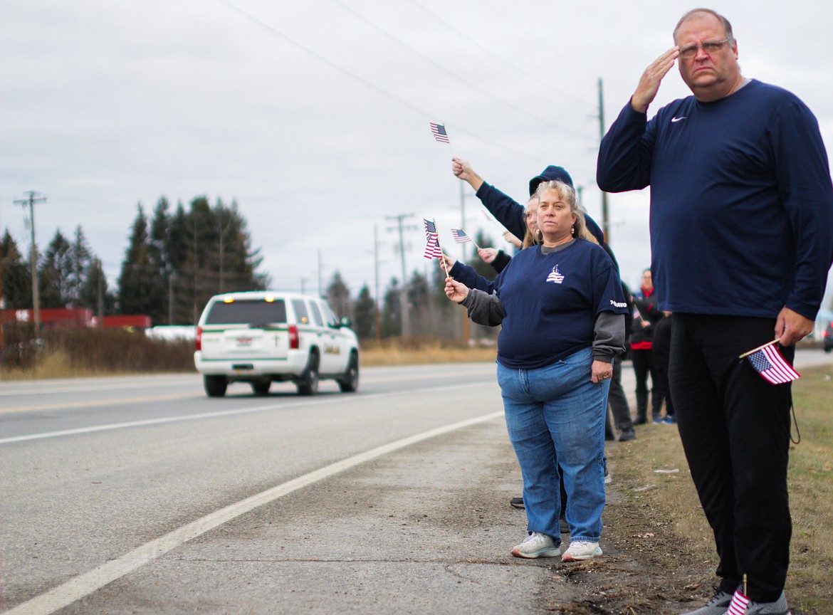 Lore Steiner (left) stands with a small crowd by the roadside watching the procession of first responder vehicles Thursday afternoon on Highway 200.