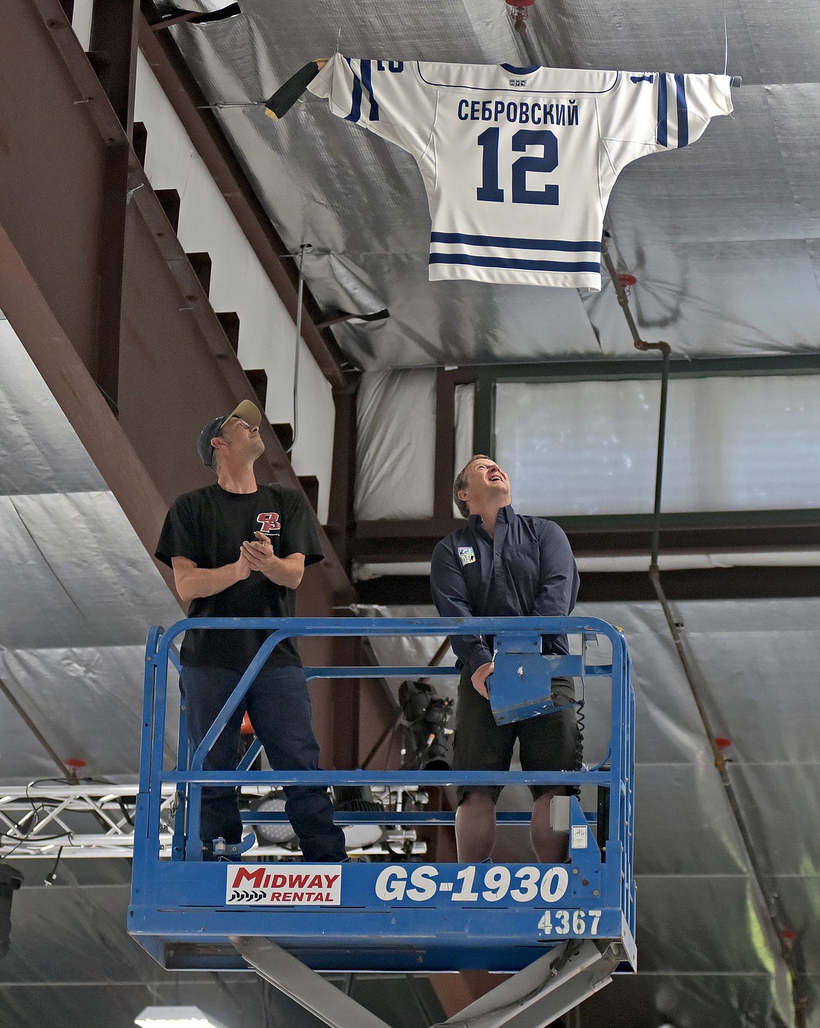 Stumptown Ice Den General Manager Greg Harms, right, and rink worker Mike Lenz, left, hang local hockey advocate Ken Sebrowsky's favorite jersey in the rafters of the ice rink on Thursday, Jun. 3. (Whitney England/Whitefish Pilot)