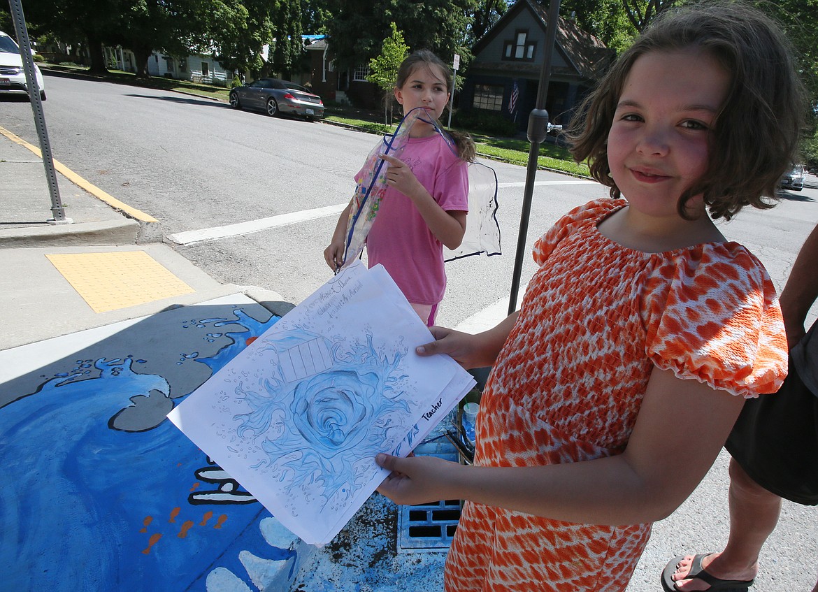 Incoming Sorensen Magnet School fifth-grader Francesca Drake shows off concept art created by classmate Thomas Taylor as she and Madeleine O'Dowd, background, finish a storm drain mural at the school Monday.