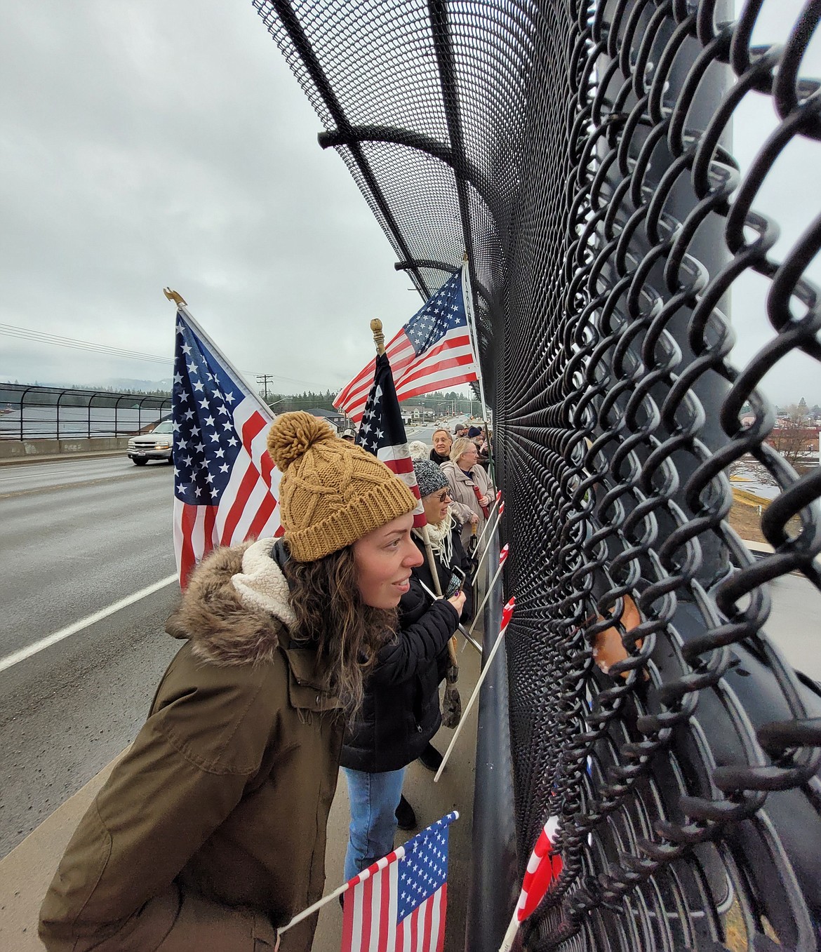 Hannah Smith of Post Falls peeks through overpass fencing Wednesday morning as she and masses of North Idahoans wave flags for the American Freedom Convoy.