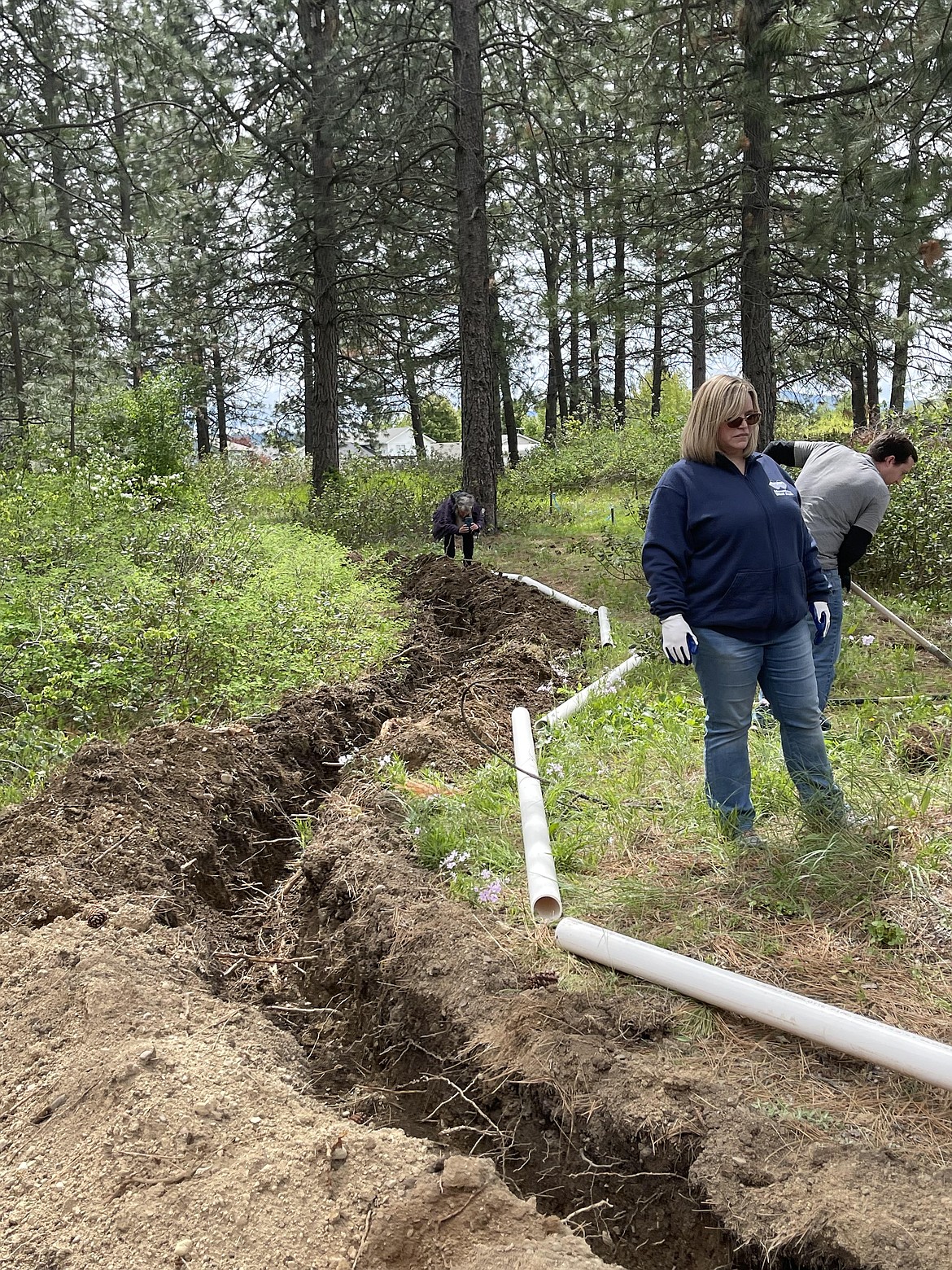 Volunteers stand next to the 100 foot trench dug at the Children's Village sensory park Friday. In the foreground are Mountain West Bank volunteers Shawna Davidson and Tyler Lohman. Lohman traveled all the way from Boise to help with the project. A pipe was placed in the trench Friday afternoon and will connect the two ends of a speaker phone. Children on each end will be able to communicate with each other from across the wooded area.