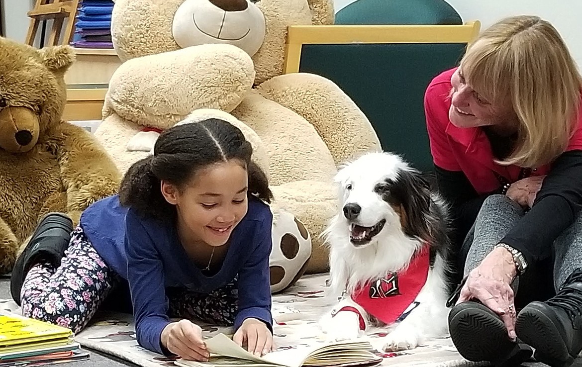 A girl reads to therapy dog Ande, with Ande's team member Susie Gonaver assisting.