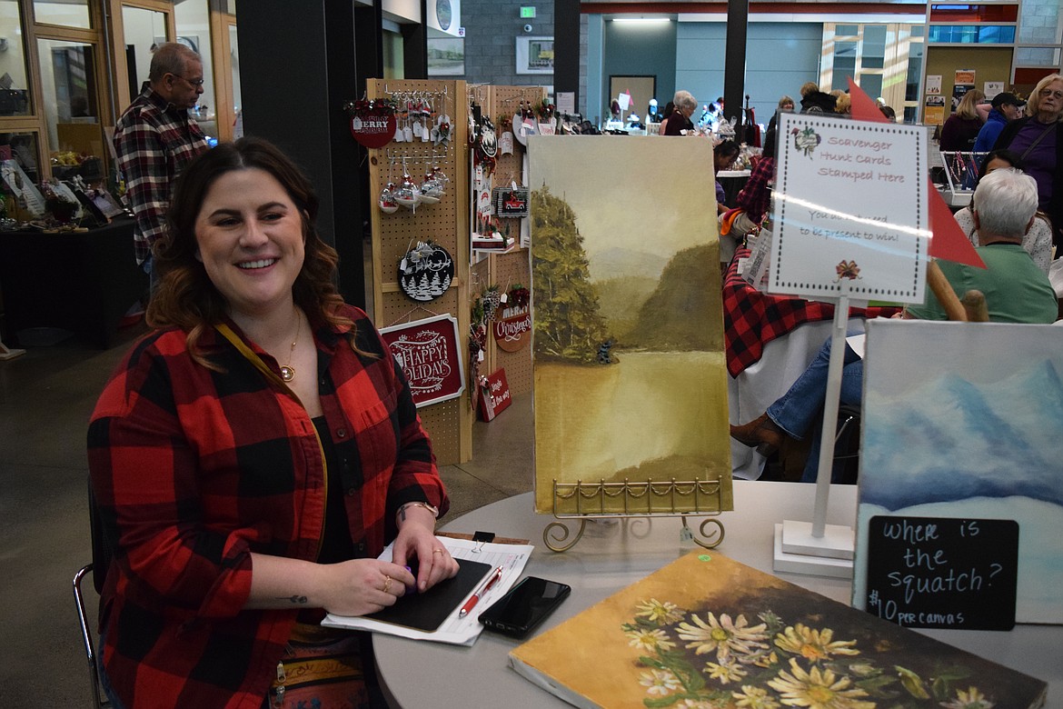 Christie Stensland of the Coulee City Art Club sits at the annual Holiday Bazaar at the Columbia Basin Technical Skills Center on Saturday with paintings the club has altered with stickers of Sasquatch. “It’s fun,” she said, and something the club does with older, unsold paintings to help raise money.