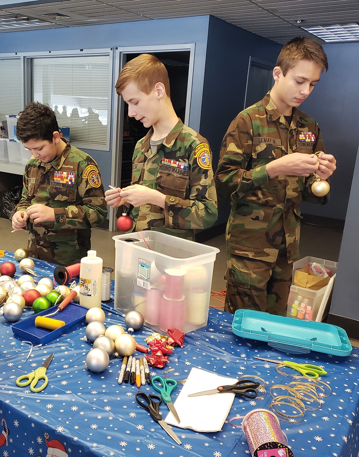 Young Marines volunteers (from left) Lance Cpl. Anthony Fox, Pfc. Daymian Reese, and Pfc. Michael Lemay string ornaments to be painted with thumbprint reindeer or handprint snowmen as kids return from shopping. Volunteering for the Holidays and Heroes event aligns with the Young Marines' goals of teaching leadership, teamwork and discipline.