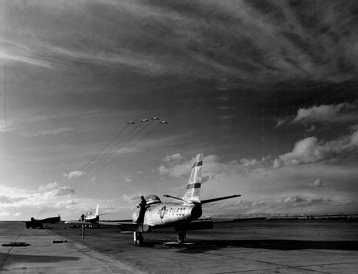 Four F-86 fighter jets, likely with the 71st Fighter Squadron, fly over a pair of jets sitting on the runway of Larson Air Force Base sometime in the summer of 1950. The view is looking west toward Ephrata.