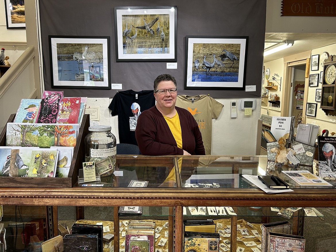 Gallery Director Jenn Stevenson sits at the front desk of the Old Hotel Art Gallery in Othello, surrounded by a case of jewelry, postcards, information, and some art by Sandy Taggares in the background.