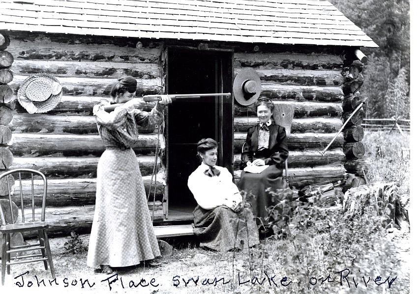 A woman shooting a gun, next to two other women seated in front of a cabin at the Johnson Place near the Swan River, circa 1900. It is now the Kootenai Lodge. (courtesy of the Northwest Montana History Museum)