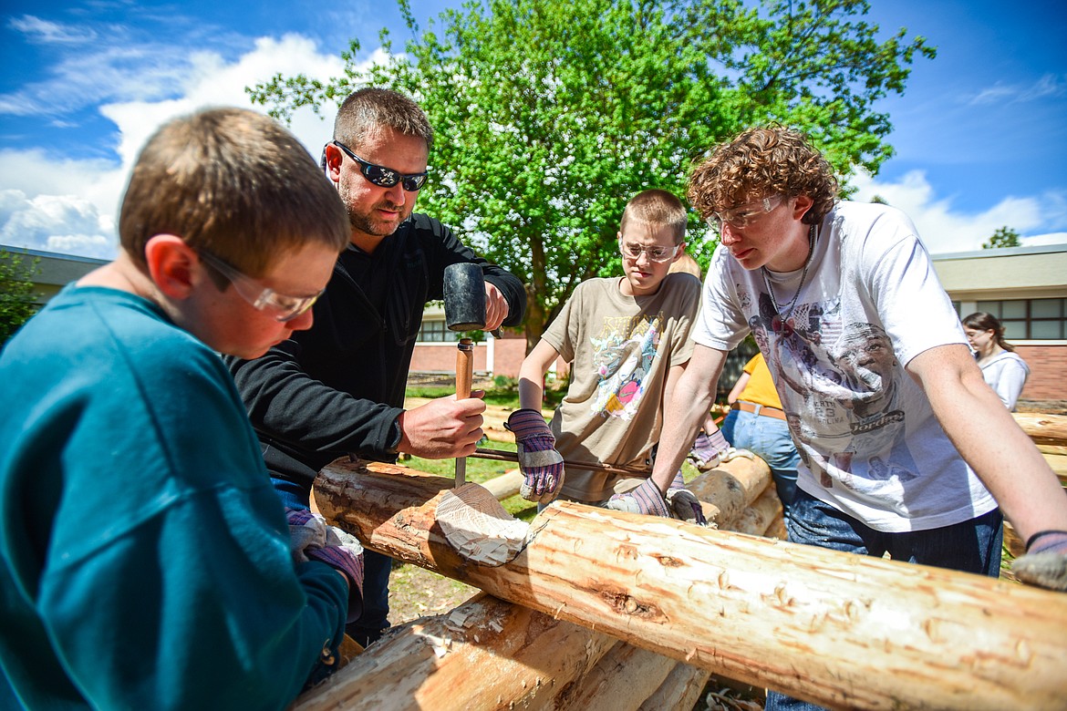 Kalispell Middle School teacher Kris Schreiner helps, from left, Rowan Wisher, Gavin Baker and Cooper Pelc cut a saddle notch as Schreiner's eighth-grade history students construct a log cabin on Tuesday, May 9. (Casey Kreider/Daily Inter Lake)