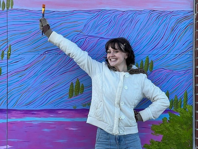 Emma Buchanan, a graduate of Big Bend Community College’s art program and a Soap Lake resident of three years, holds a paintbrush in front of her mural in Soap Lake.