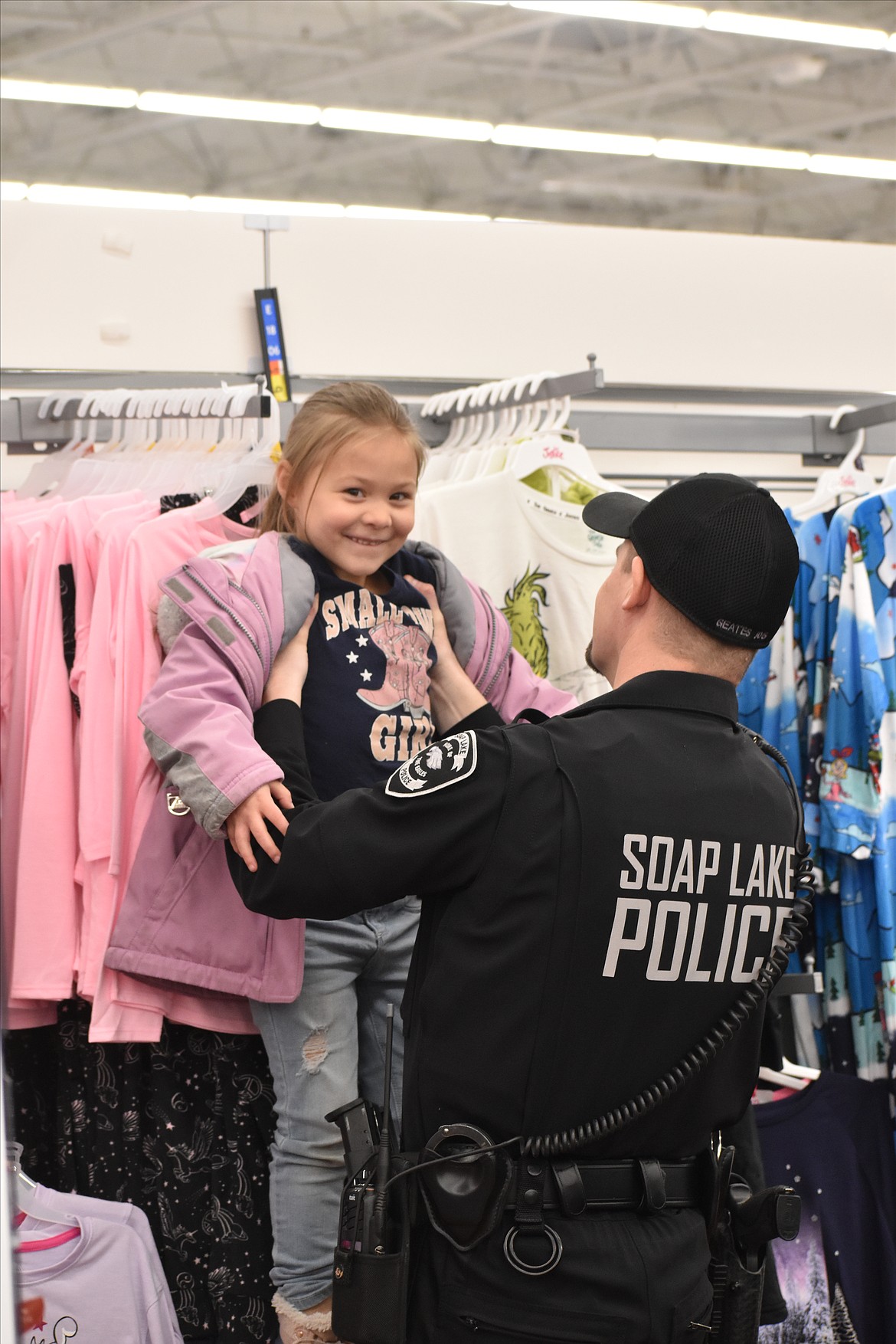 A girl smiles as Soap Lake Police Officer Robert Geates holds her up to see if she would fit any of the clothes on the rack at the 2023 Shop With a Cop event.