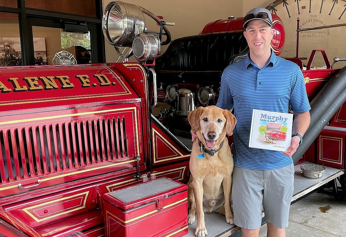 Coeur d'Alene firefighter Cody Moore, joined by Murphy at the Coeur d'Alene fire station, holds a copy of his book "Murphy Gets A Job."