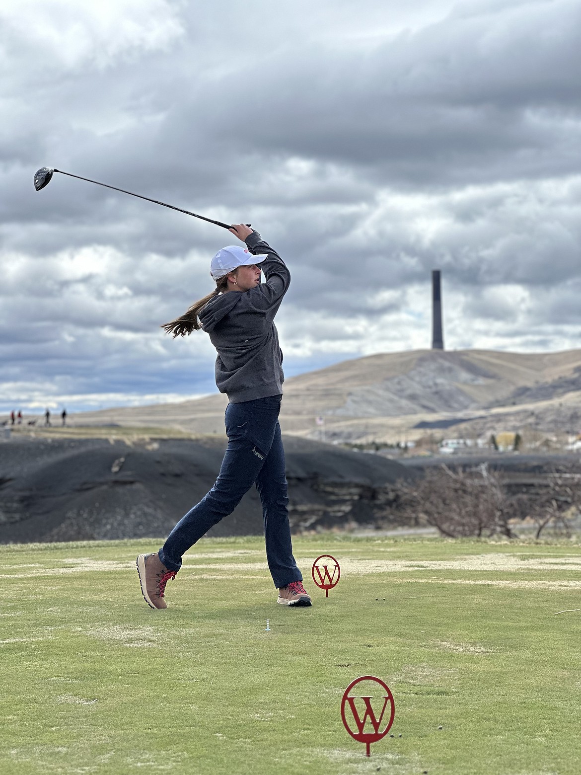 With the omnipresent Anaconda copper smelter smokestack standing in the background, T Falls freshman golfer Aubrey Baxter tees off during last week's Western Division golf tourney under cold and windy conditions.  (Photo by Doree Thilmony)