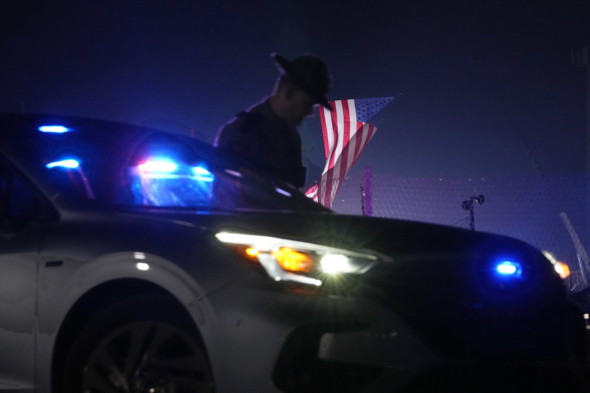 An officer stands at his car early Sunday, July 14, 2024, closing access to the site of the rally where former President Donald Trump was the target of an assassination attempt on Saturday in Butler, Pa.. Behind the officer, a large flag from the rally still flies. (AP Photo/Sue Ogrocki)