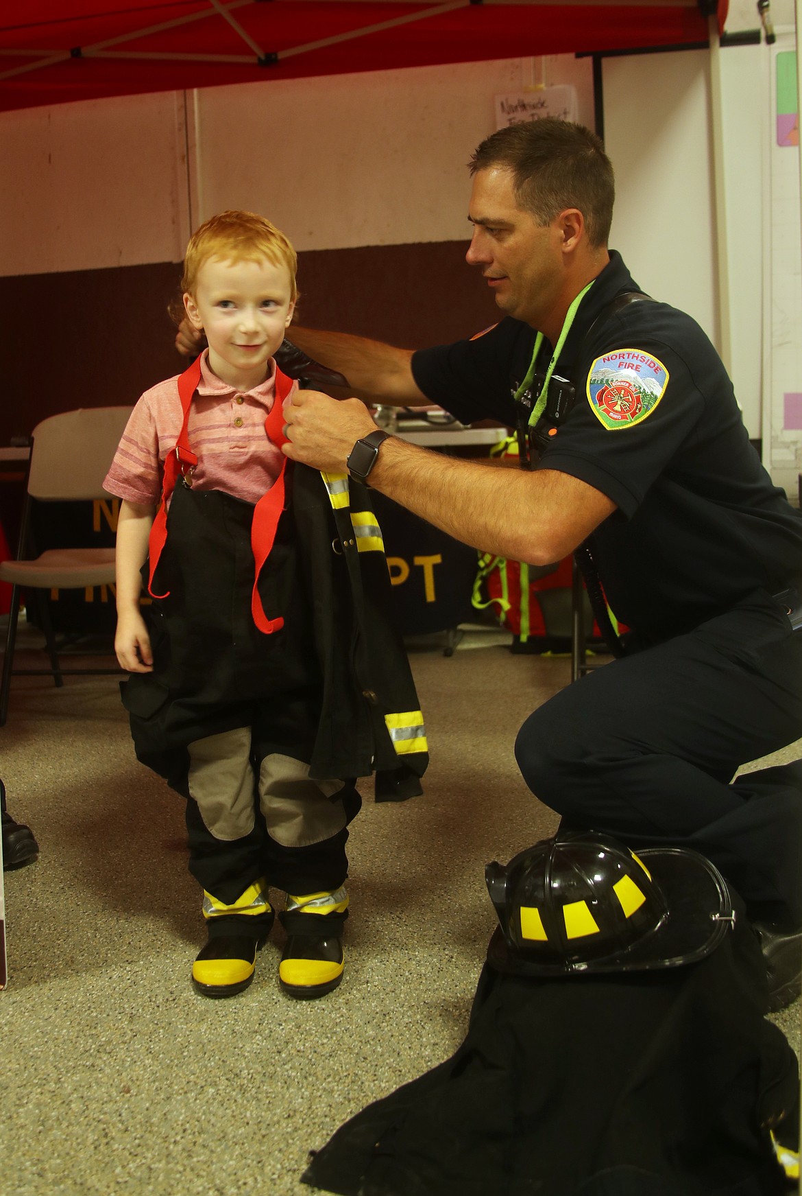 Fynn Morgan, the son of Ashley Offermann and Troy Morgan gets fitted for a pair of turnouts by a Selkirk Fire firefighter after stopping by the district's booth at the Bonner County Fair on Wednesday.