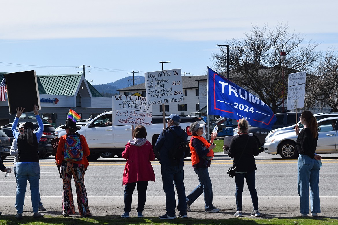 People protesting the Trump administration hold signs along U.S. 95 in Coeur d'Alene on Saturday as a motorist drives by flying a 'Trump 2024' flag.